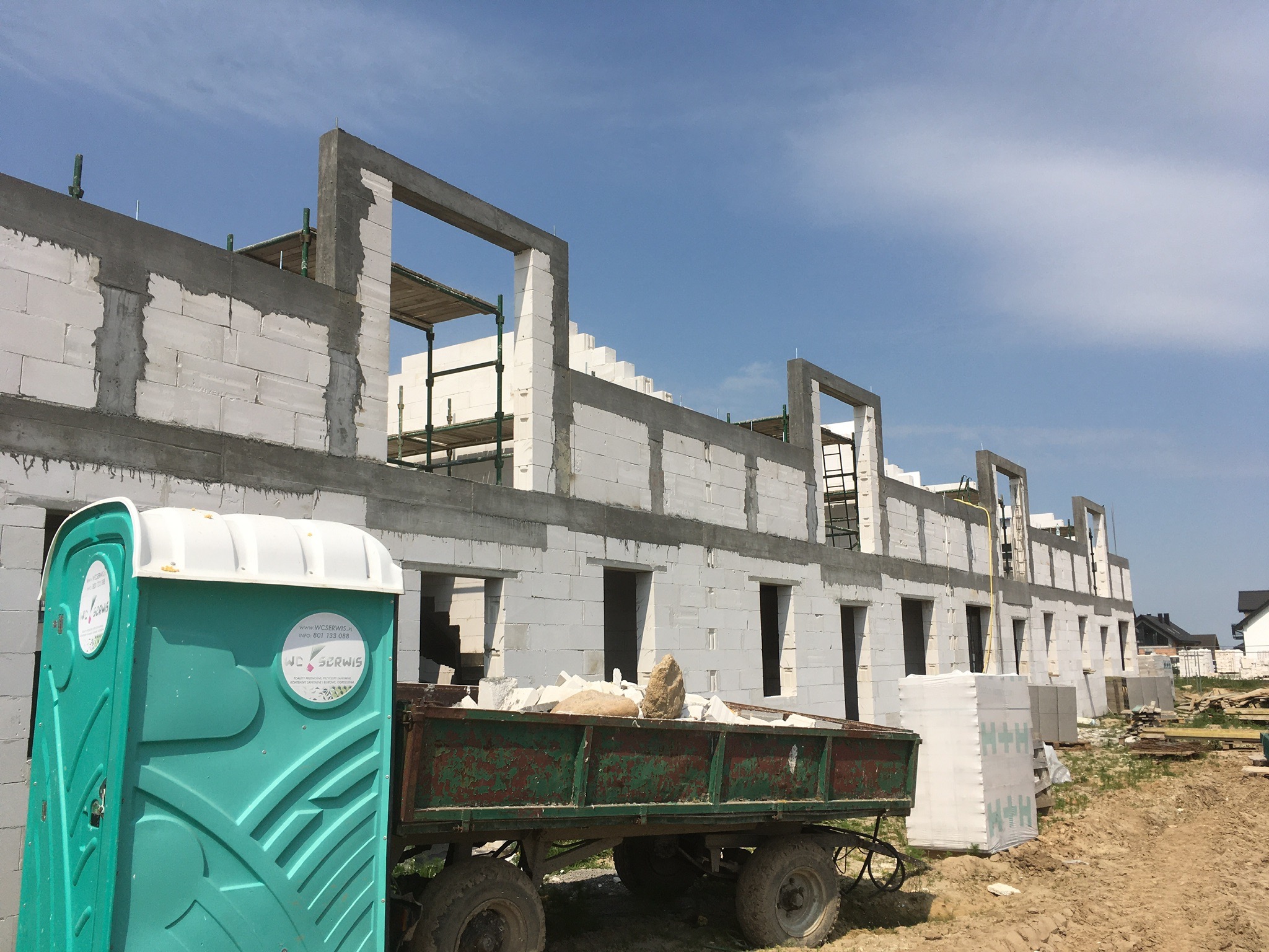 Wznoszenie ścian domu z bloczków betonu komórkowego Ytong, widoczny fragment rusztowania, przyczepa z gruzem i przenośna toaleta na placu budowy.