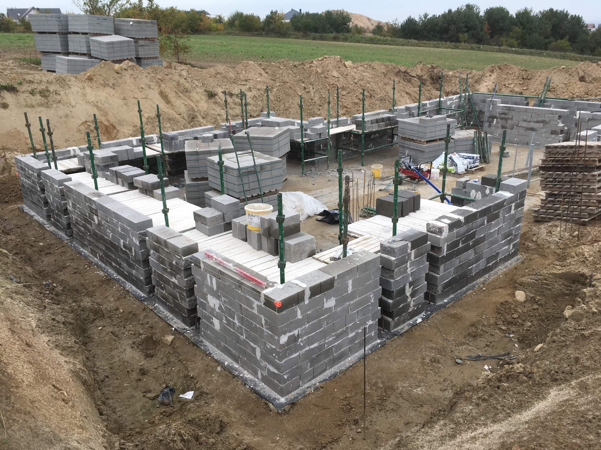 Wznoszenie ścian fundamentowych z bloczków betonowych na placu budowy, widoczne podpory szalunkowe i palety bloczków w tle.