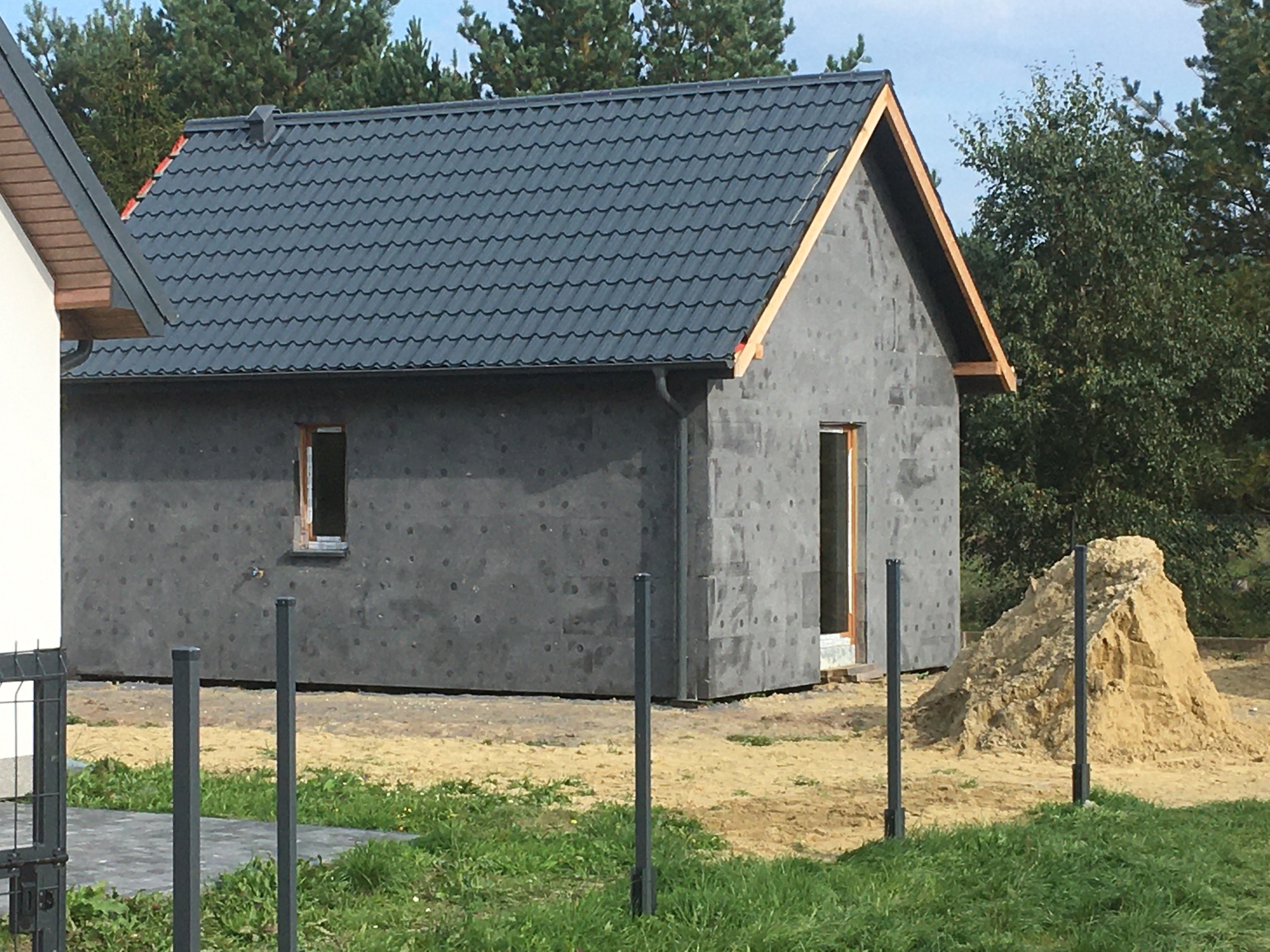 Dom w trakcie budowy: szara elewacja z widocznymi otworami, czarny dach z metalowej dachówki, widoczne drewniane elementy konstrukcji dachu, hałda piasku obok budynku i fragment ogrodzenia.