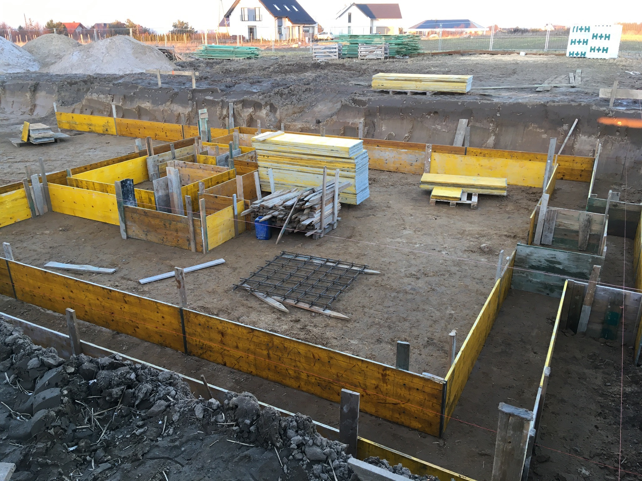Wykonanie szalunków pod fundamenty domu jednorodzinnego z widoczną siatką zbrojeniową, deskami, stertą materiałów budowlanych i wykopem w tle.