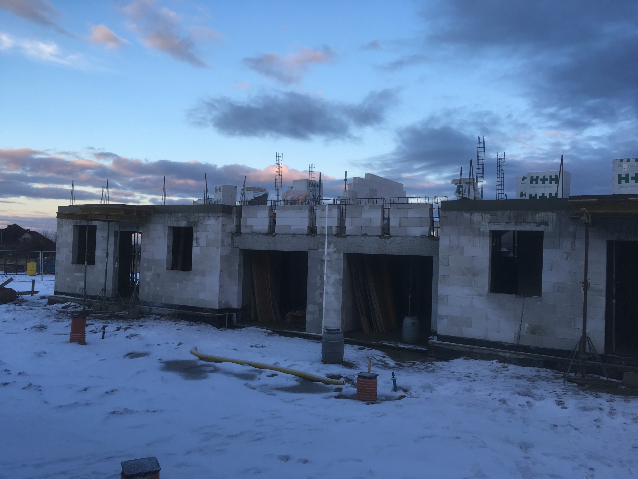 Surowy stan domu w trakcie budowy z bloczków betonu komórkowego w zimowej scenerii, widoczne zbrojenia i materiały budowlane, tło stanowi pochmurne niebo o zmierzchu.