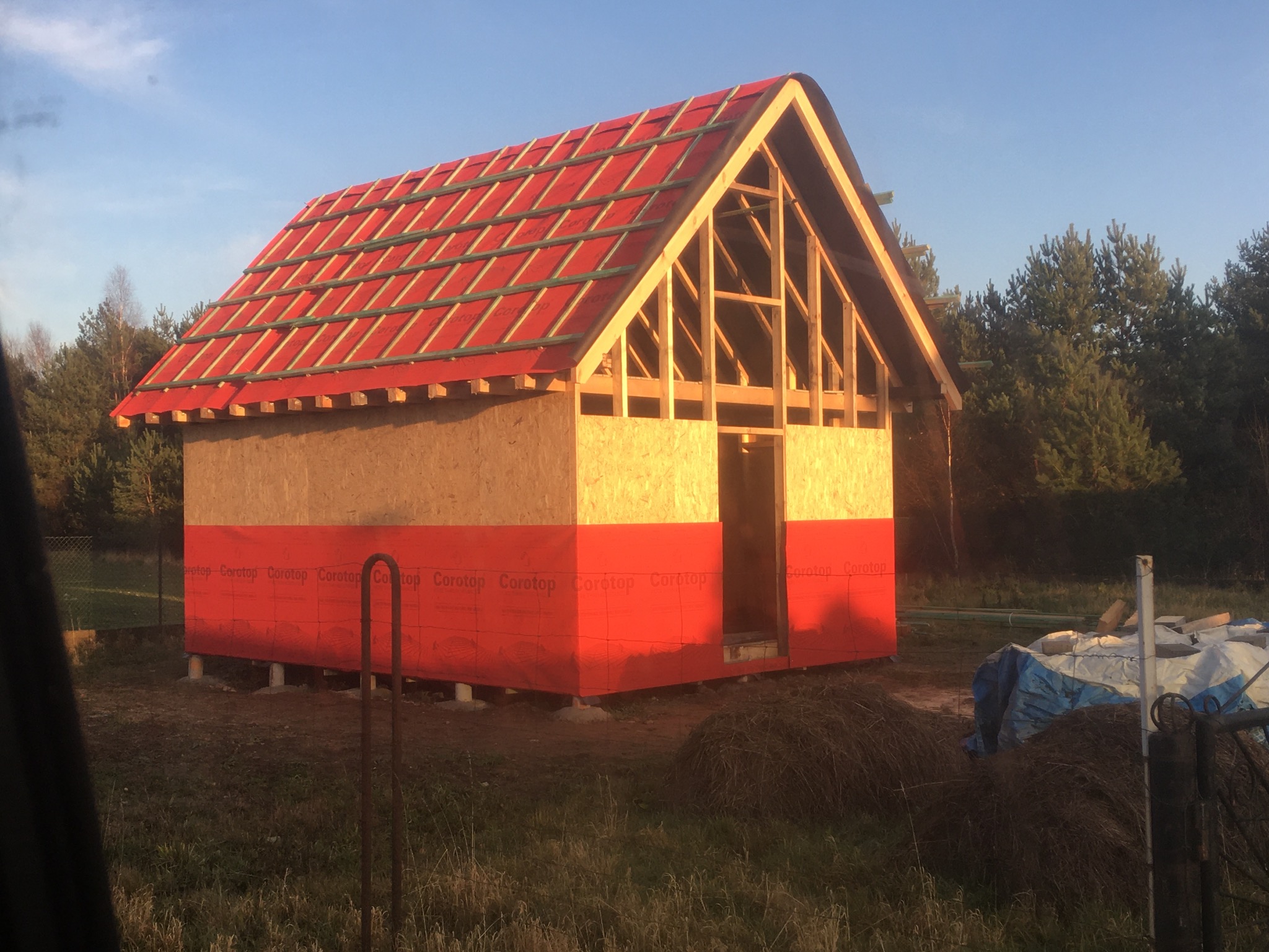 Niedokończony szkielet domu z deskami OSB i czerwoną membraną na dolnej części ścian, widoczne krokwie i częściowo położone czerwone pokrycie dachu z łatami, na zewnątrz sterta siana i plandeka.
