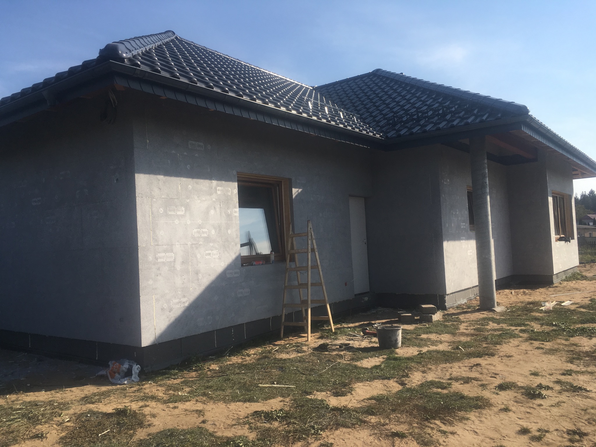 Surowy stan domu jednorodzinnego z szarą elewacją i ciemną dachówką, widoczna drabina oparta o ścianę, teren wokół domu częściowo zagospodarowany, niebo błękitne.