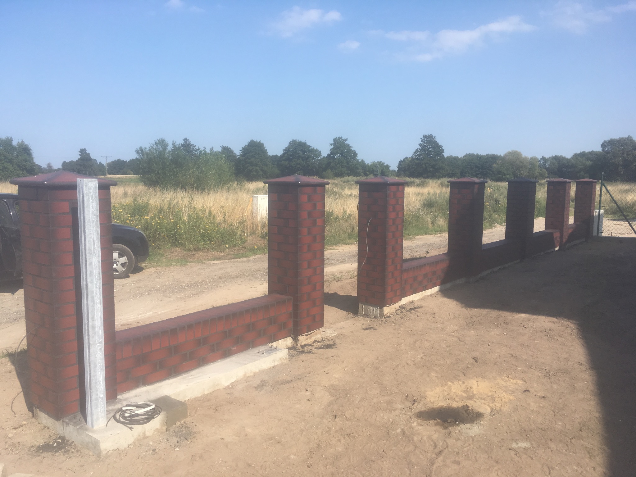 Częściowo wzniesione ogrodzenie z cegły klinkierowej z metalowym słupkiem, widoczne przewody elektryczne, w tle pole i drzewa.