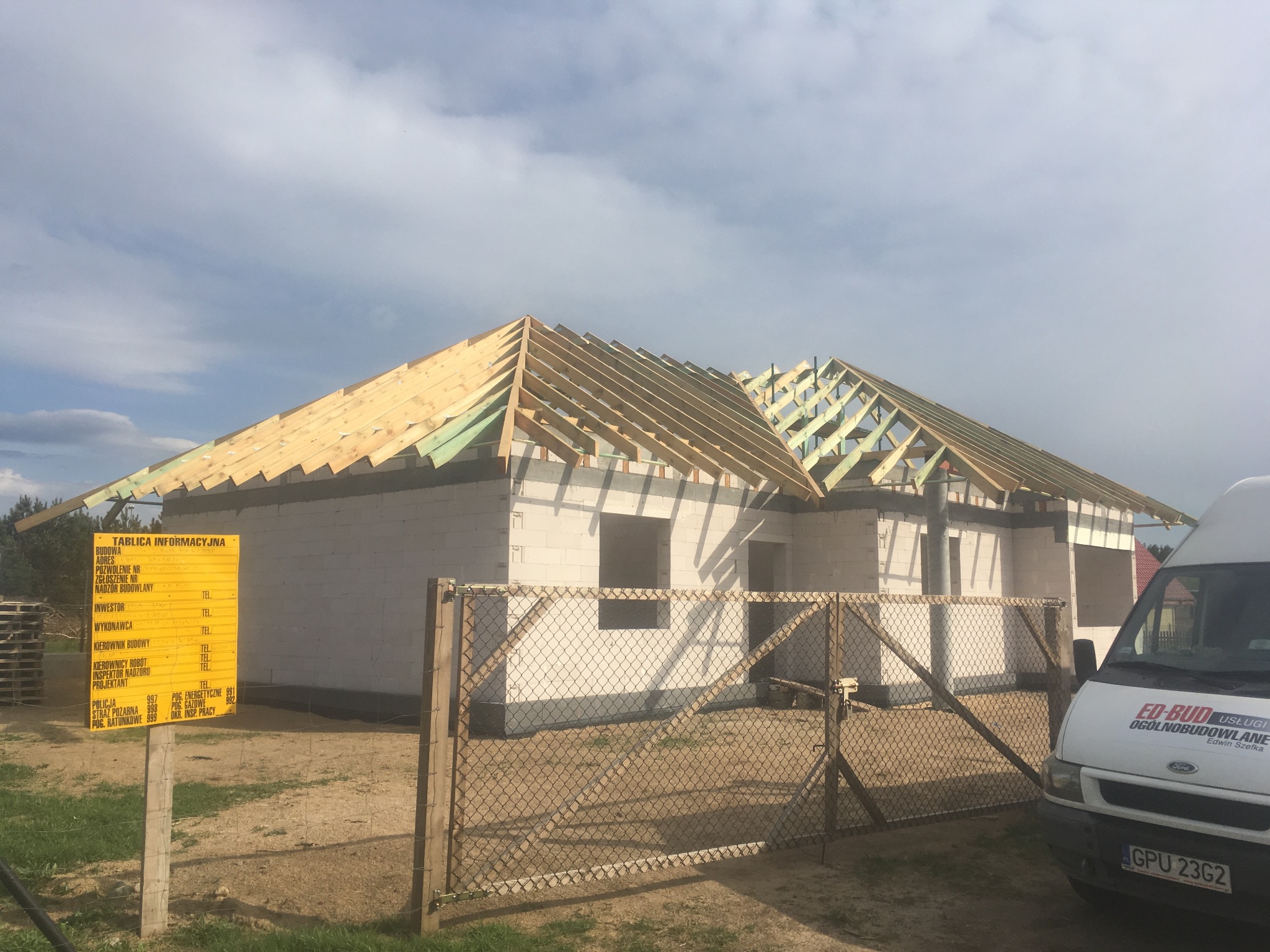 Surowy stan domu z białego bloczka, widoczna konstrukcja dachu z drewnianych belek, tablica informacyjna budowy i samochód firmy budowlanej z logo.