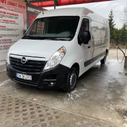 Biały Opel Master w myjni samochodowej, z widocznymi śladami piany na karoserii i kostce brukowej pod kołami.