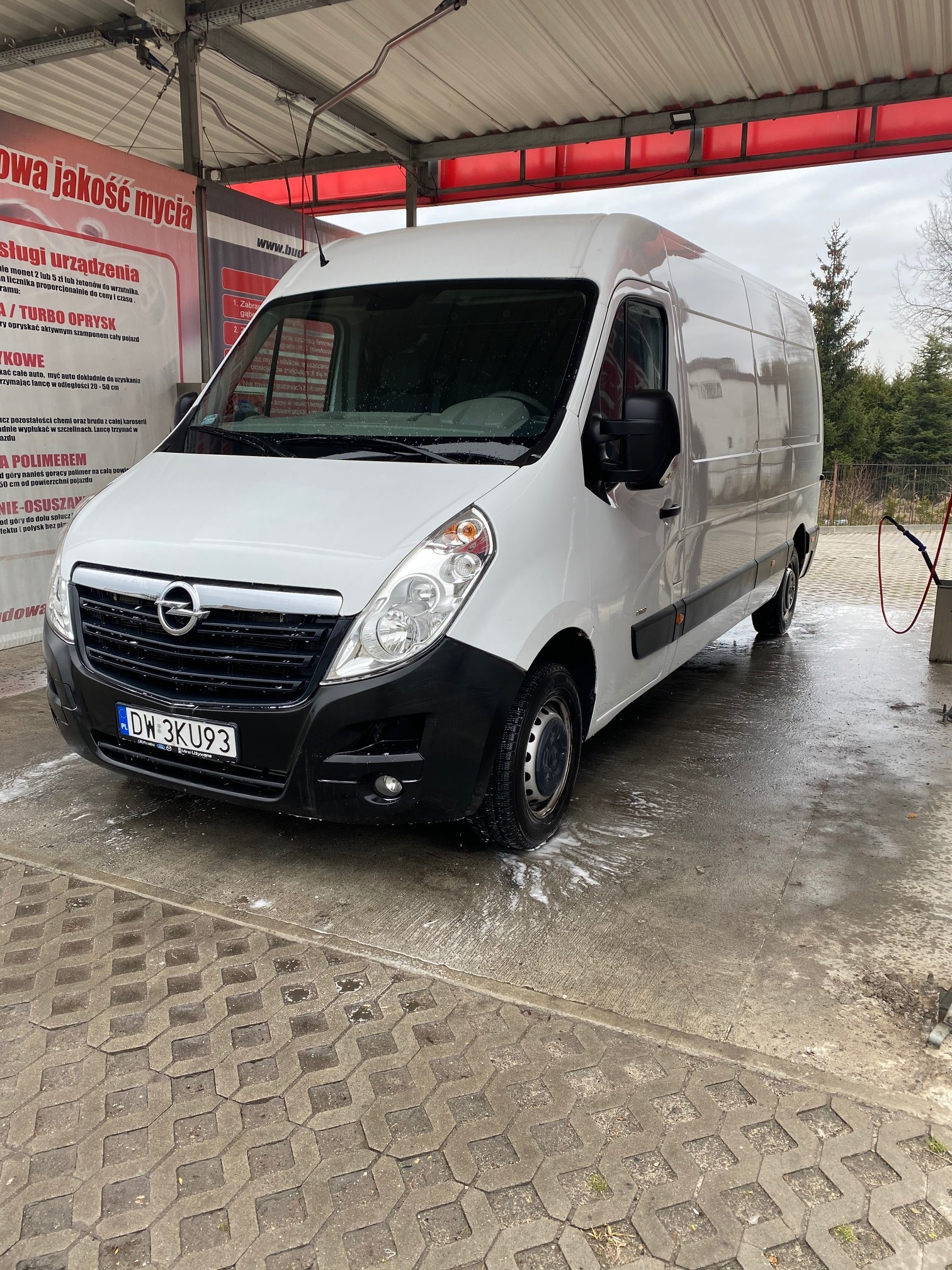 Biały Opel Master w myjni samochodowej, z widocznymi śladami piany na karoserii i kostce brukowej pod kołami.
