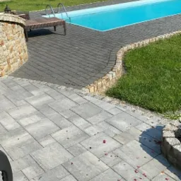 Szara kostka brukowa ułożona wokół basenu w Bielsku-Białej, widoczne drobne kwiatowe płatki na powierzchni. W tle zielony trawnik i żywopłot.