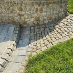 Obrzeże rabaty z kostki granitowej, schody z płyt betonowych i trawnik, całość oświetlona przez lampę ogrodową w stylu retro.