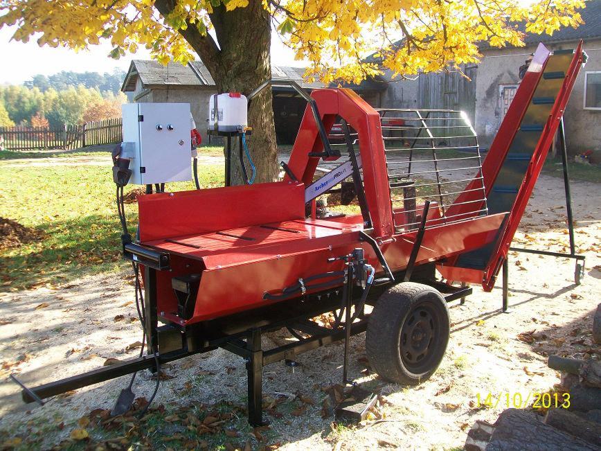 Czerwona pilarko-łuparka z podajnikiem taśmowym na przyczepie, ustawiona na zewnątrz pod drzewem z żółtymi liśćmi, widoczny panel sterowania i zbiornik na olej.