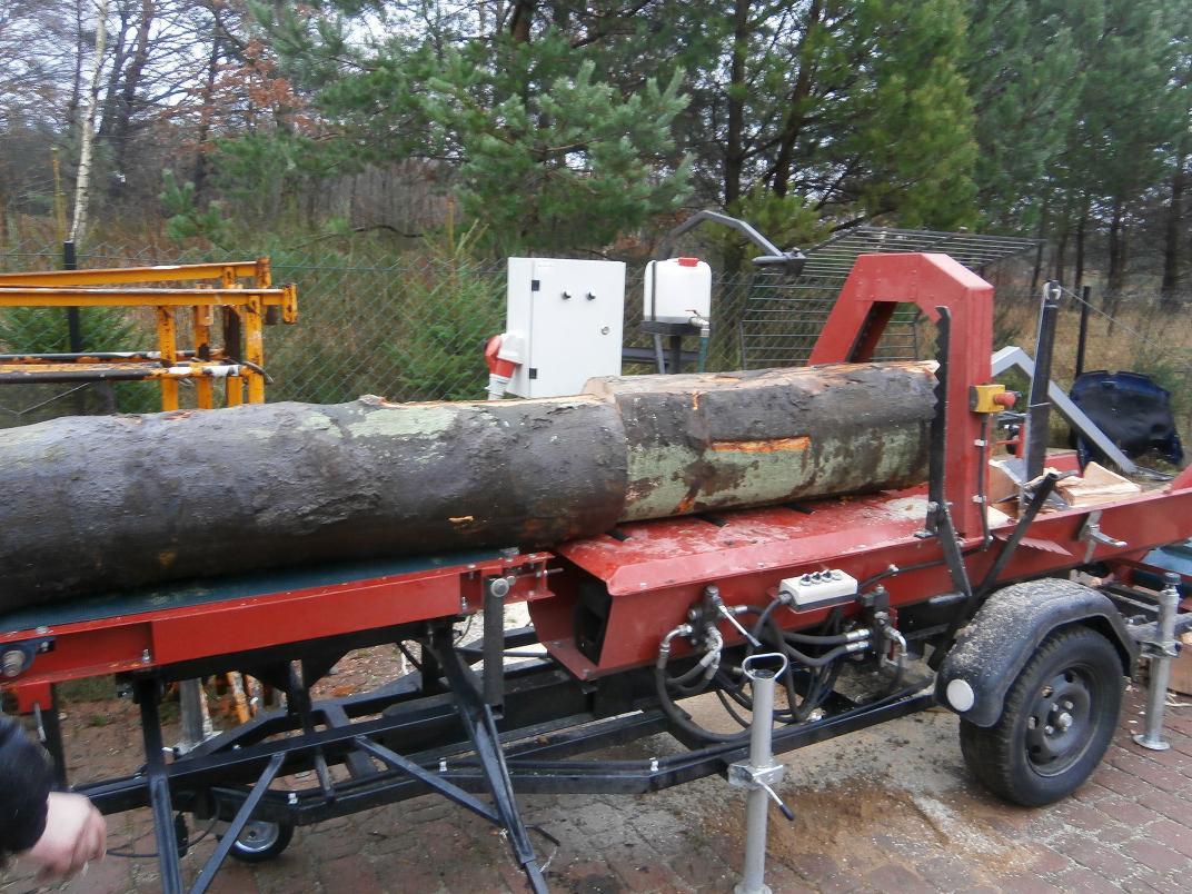 Duża kłoda umieszczona na czerwonej, mobilnej pilarko-łuparce z hydraulicznym systemem, widoczne drzewa w tle.