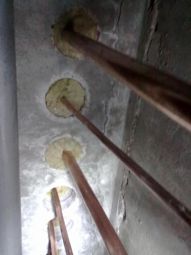 Przejścia rur przez ścianę zabezpieczone wełną mineralną, widoczne ślady po wierceniu otworów. Instalacja w szarym pomieszczeniu z surowymi ścianami.