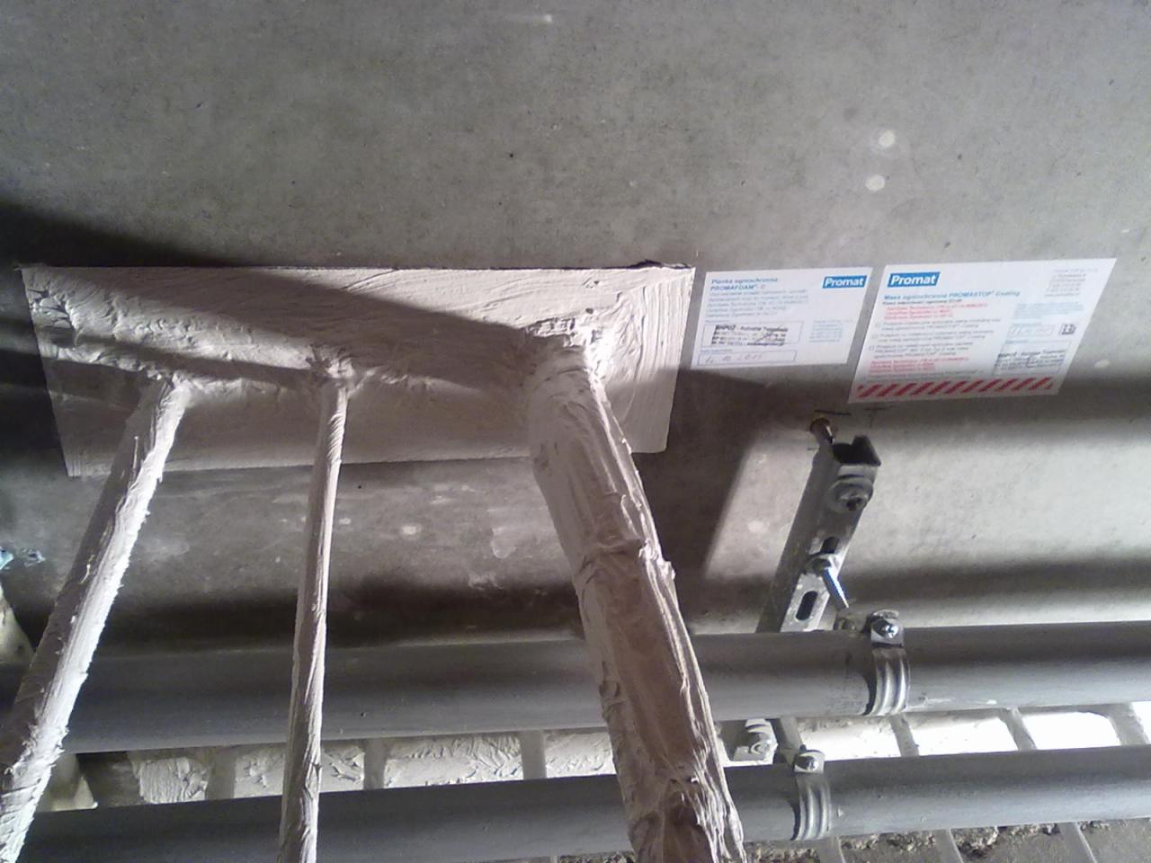 Zabezpieczone ogniochronnie rury pod stropem, pokryte białą masą. Widoczne etykiety informacyjne producenta Promat. Metalowe uchwyty mocujące rury.