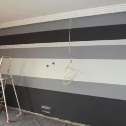 Ściana w pokoju pomalowana w poziome pasy w odcieniach szarości i bieli, widoczna drabina aluminiowa, wystające kable elektryczne ze ściany, listwa dekoracyjna pod sufitem.