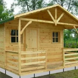 Surowa, drewniana konstrukcja małego domku z zadaszonym wejściem, widoczne elementy więźby dachowej i prostokątne okna z podziałami, całość na tle zieleni.