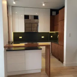 Nowoczesna kuchnia z białymi frontami szafek, czarnym blatem i drewnianymi akcentami, oświetlona ledowym paskiem pod szafkami wiszącymi, z barkiem oddzielającym przestrzeń.