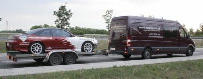 Czerwono-biały samochód sportowy przewożony na przyczepie przez ciemnobrązowego busa na poboczu drogi.