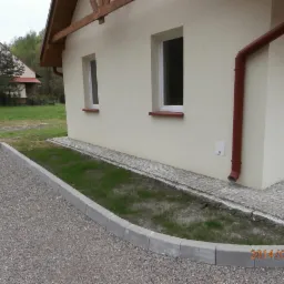 Wykończony teren wokół domu z szarą kostką brukową, obrzeżami i drobnym żwirem. Widoczne okna i rynna na elewacji budynku w tle.