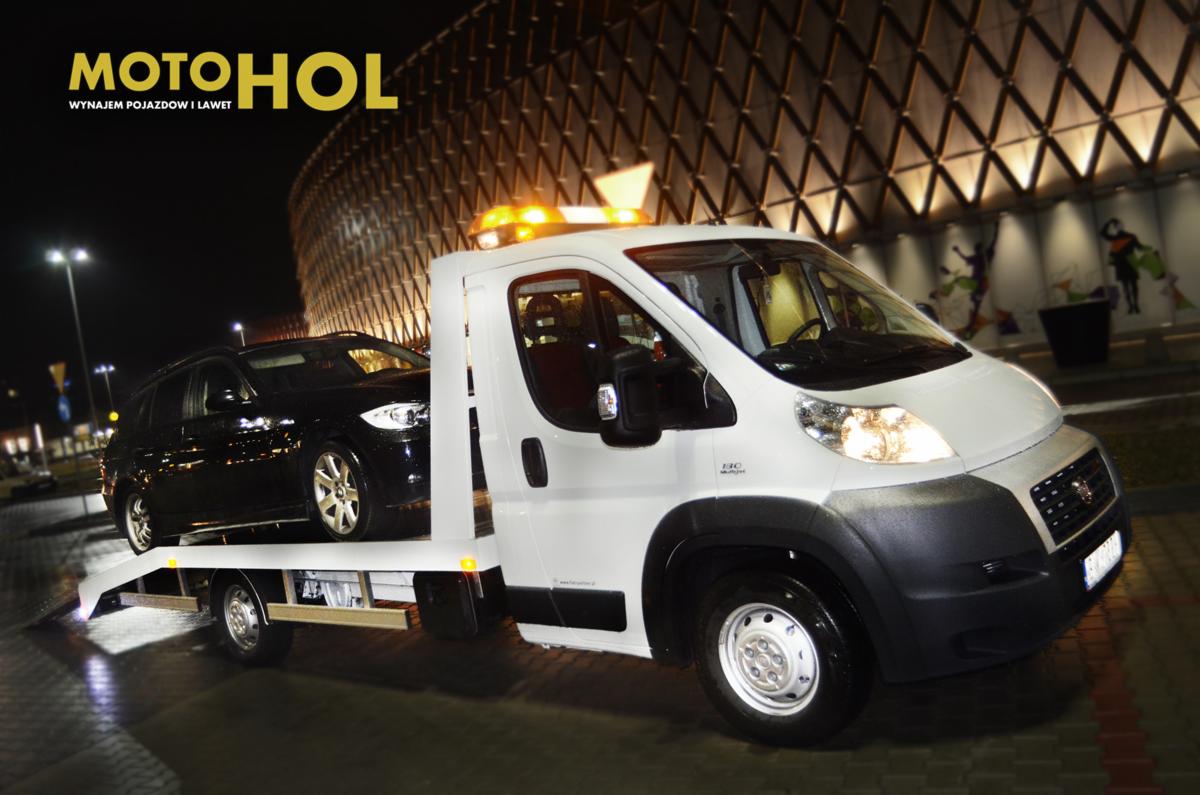 Biała laweta Fiat Ducato z włączonymi światłami ostrzegawczymi przewozi czarny samochód osobowy na tle nowoczesnego budynku oświetlonego latarniami ulicznymi.