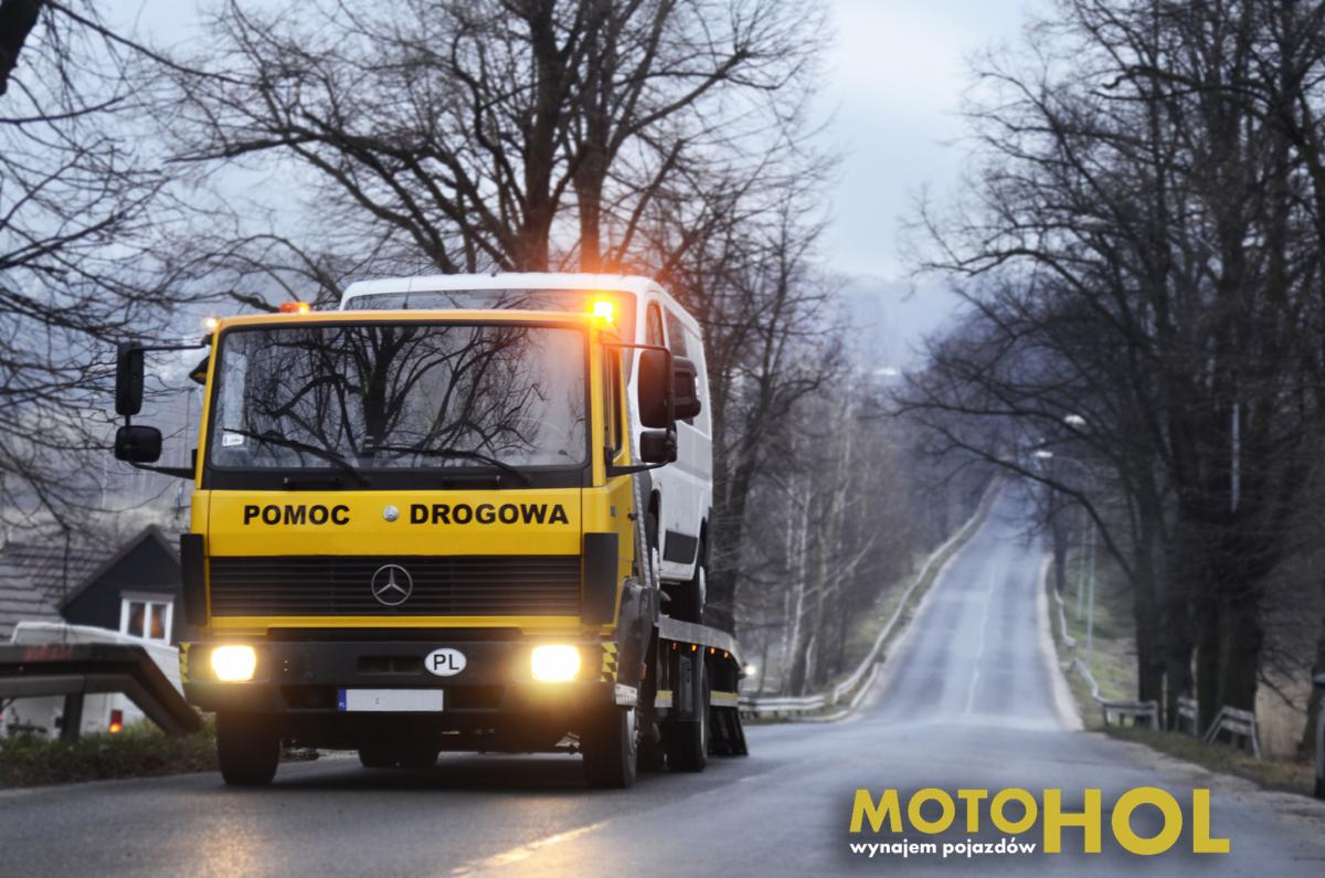 Żółty samochód pomocy drogowej z napisem 'Pomoc Drogowa' i polską tablicą rejestracyjną transportuje biały bus na lawecie, w tle widoczna droga w pochmurny dzień.