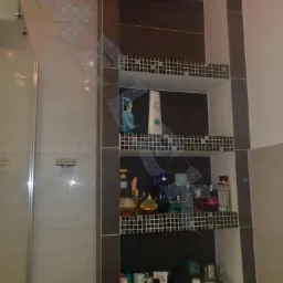 Półka wnękowa w łazience wyłożona mozaiką, z trzema poziomami wypełnionymi kosmetykami i akcesoriami, obok kabina prysznicowa z szklanymi drzwiami.