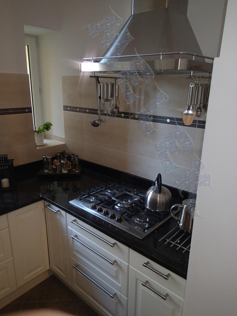 Fragment kuchni z białymi szafkami z długimi, poziomymi uchwytami, czarnym blatem, płytą gazową i metalowym okapem z relingiem na akcesoria kuchenne, wykończona beżowymi płytkami z ciemnobrązowym...