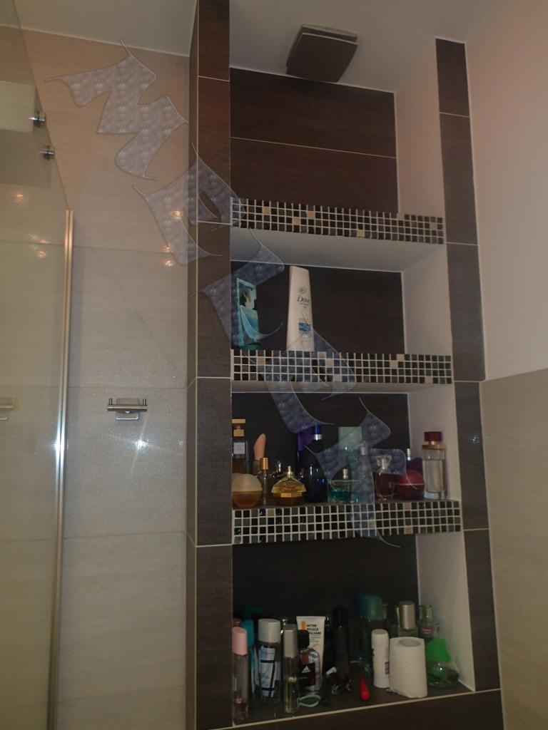 Półka wnękowa w łazience wyłożona mozaiką, z trzema poziomami wypełnionymi kosmetykami i akcesoriami, obok kabina prysznicowa z szklanymi drzwiami.