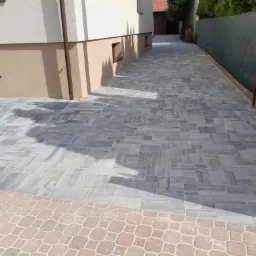 Świeżo ułożona, szara kostka brukowa na podjeździe, z widocznym fragmentem domu z beżową elewacją i ciemnymi oknami oraz ogrodzeniem z siatki w tle.