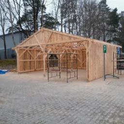 Drewniana konstrukcja w trakcie budowy w Choroszczy, widoczne elementy szkieletu dachu i ścian, teren utwardzony kostką brukową.