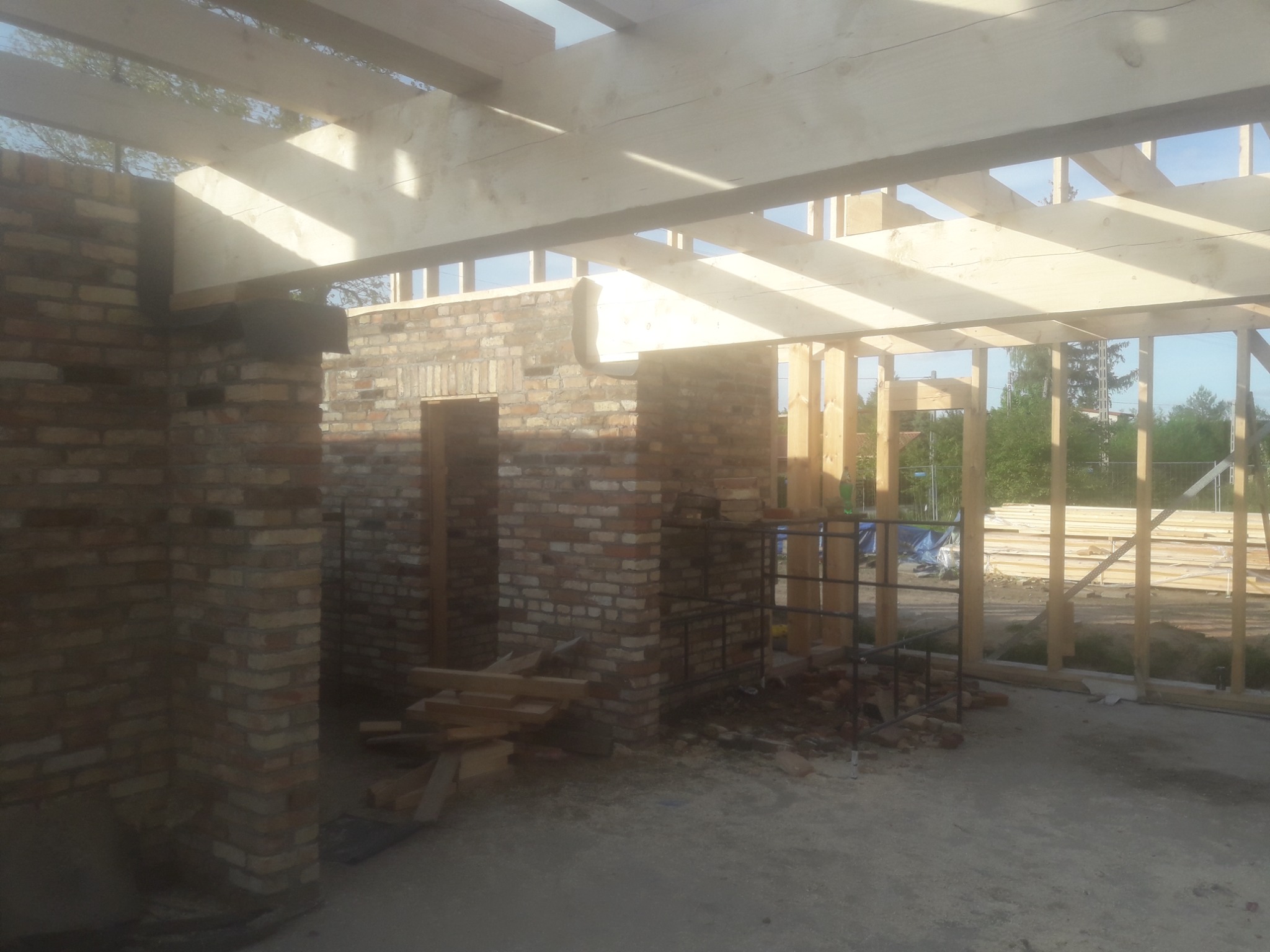 Surowy stan budynku w trakcie budowy: widoczne ceglane ściany, drewniana konstrukcja dachu oraz podłoga w stanie roboczym.