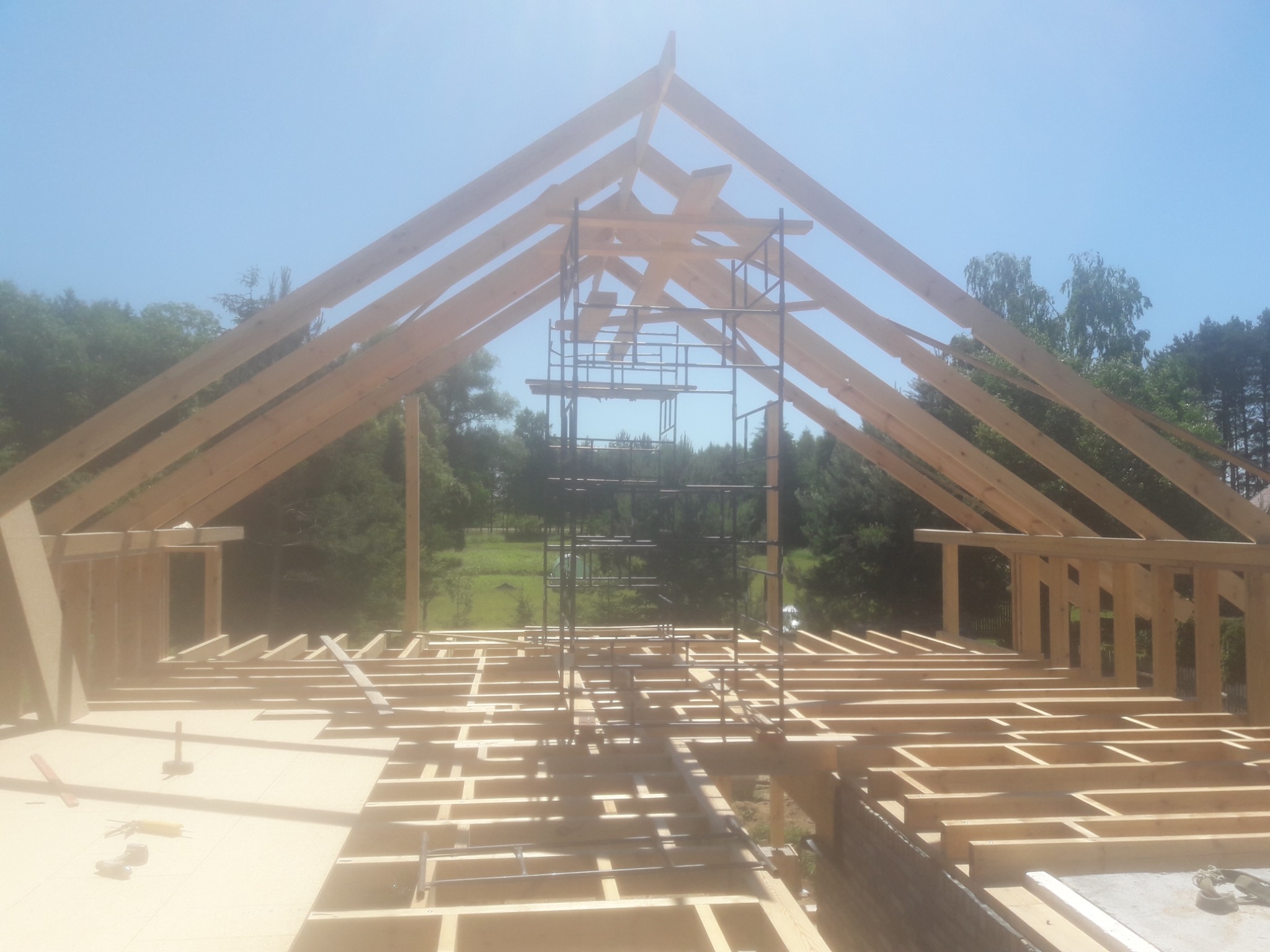 Konstrukcja drewniana dachu w trakcie budowy, widoczne elementy więźby, rusztowanie, narzędzia i fragment krajobrazu w tle.