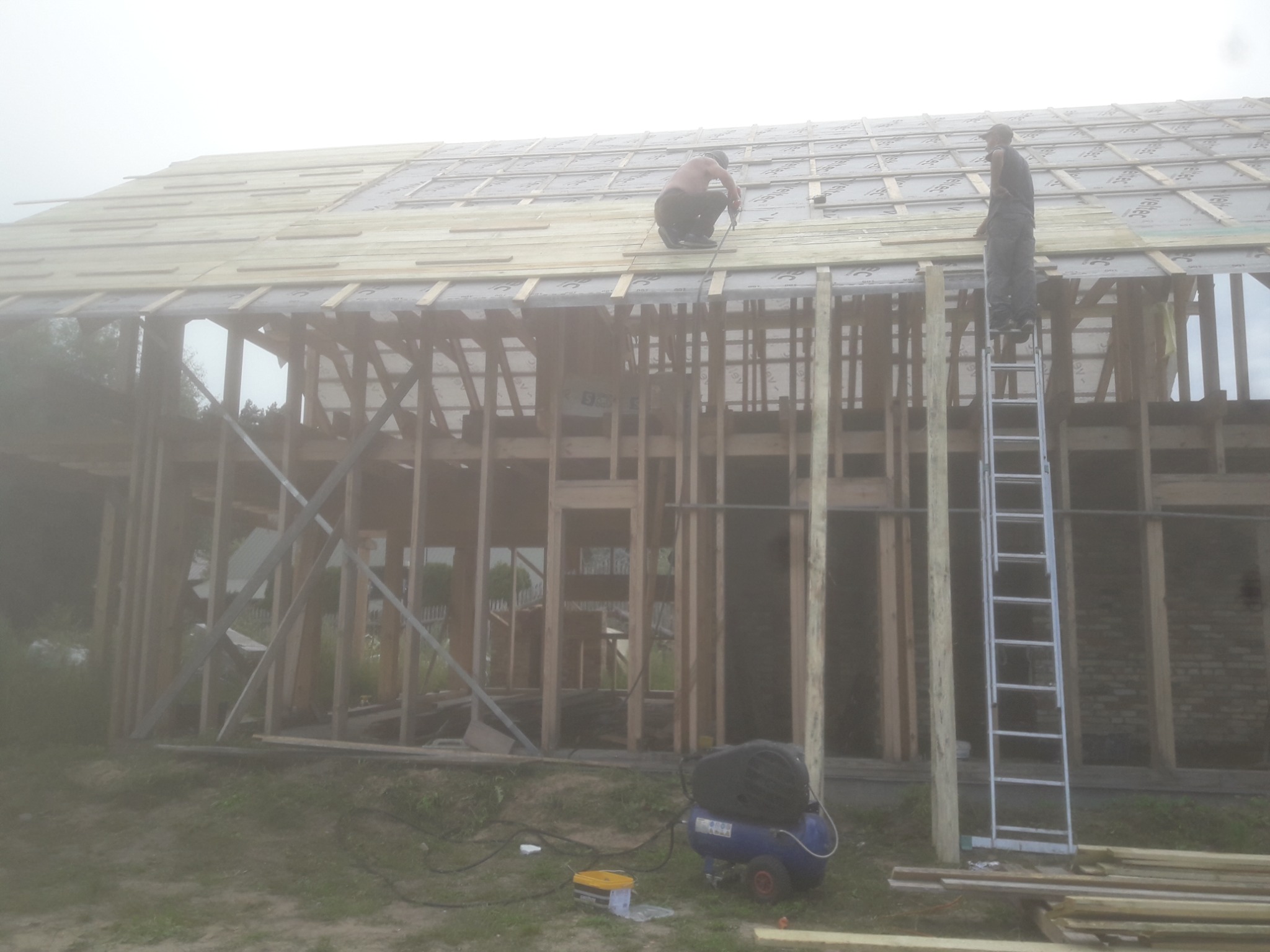 Dwóch mężczyzn pracuje na dachu szkieletu domu, jeden siedzi i przybija deski, drugi stoi na drabinie, w tle mgła i widoczna konstrukcja budynku.