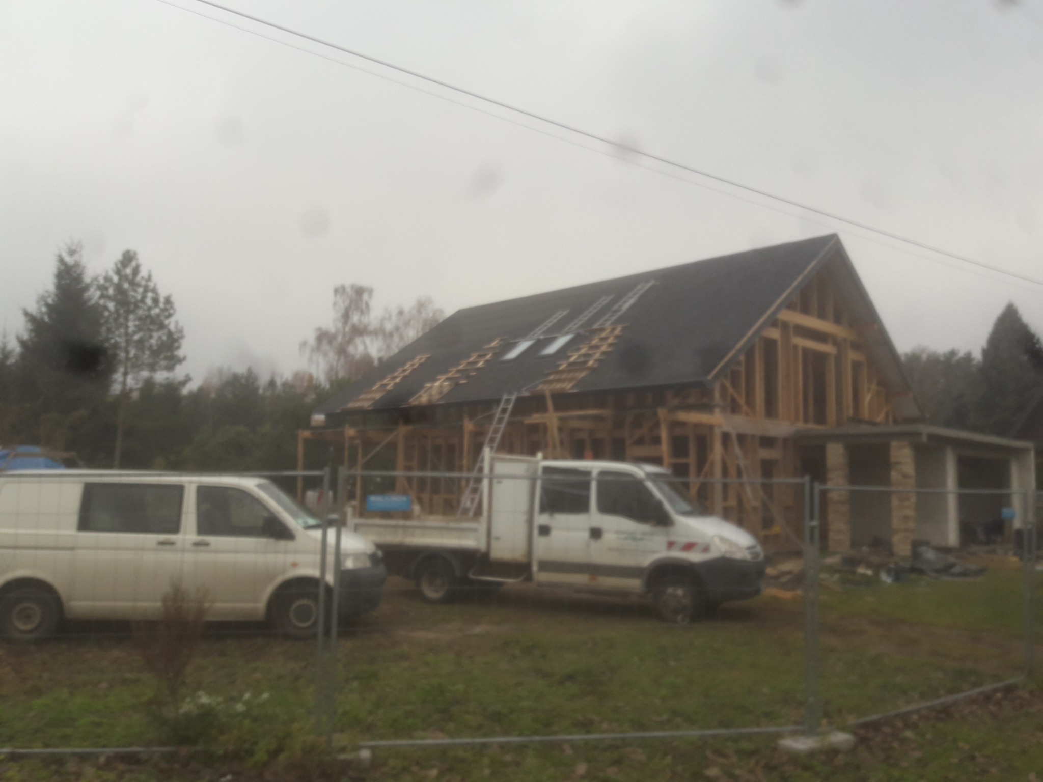 Wznoszona konstrukcja domu jednorodzinnego z widocznym szkieletem drewnianym dachu i fragmentem pokrycia, dwa samochody dostawcze na trawiastym terenie budowy w pochmurny dzień.