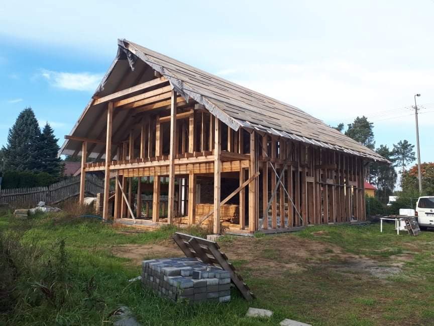 Surowy szkielet drewnianego domu w trakcie budowy, z częściowo ułożonym pokryciem dachowym, widok z zewnątrz na tle zieleni i błękitnego nieba.
