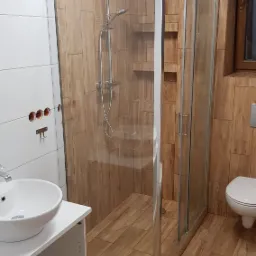 Wykończona łazienka z kabiną prysznicową typu walk-in, umywalką nablatową na białej szafce i drewnopodobnymi płytkami na ścianach i podłodze. Widoczne niedokończone otwory na ścianie po lewej stronie.
