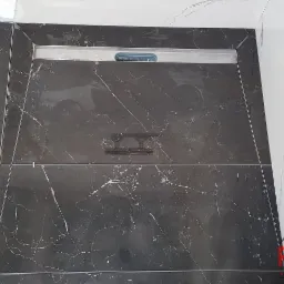 Świeżo ułożone, czarne płytki podłogowe z marmurkowym wzorem, z widocznym odpływem liniowym, częściowo zabezpieczone taśmą ochronną, kontrastujące z białymi płytkami ściennymi.