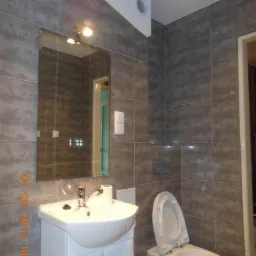 Szara łazienka po remoncie: biała umywalka z szafką, toaleta z otwartą klapą, lustro z oświetleniem. Ściany i podłoga wyłożone płytkami w odcieniach szarości.