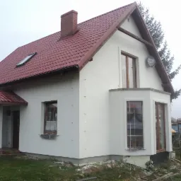 Otynkowany, biały dom jednorodzinny z czerwonym dachem, widok z boku, z widoczną anteną satelitarną i fragmentami niezabezpieczonej elewacji przy oknach.