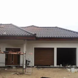 Dwukondygnacyjny dom z szarą dachówką, widoczne rusztowanie, drabiny i pracownik na dachu, teren przed budynkiem w trakcie prac wykończeniowych.