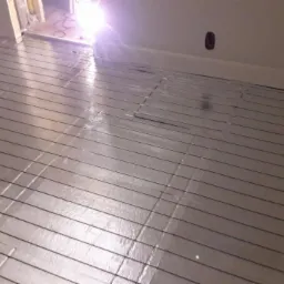 Rozłożona mata grzewcza na podłodze w pomieszczeniu z widocznym otworem w ścianie i puszką elektryczną na ścianie, przygotowana do zalania wylewką.