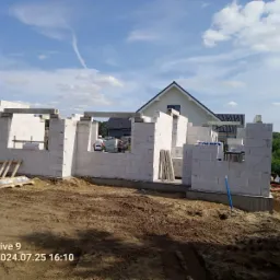 Wznoszenie ścian parteru z białych bloczk&oacute;w betonu kom&oacute;rkowego na fundamencie, widoczny pracownik w kasku, w tle dom jednorodzinny i błękitne niebo.