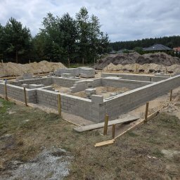 Usługi budowlane inż. Adam Andrzejewski - Częściowo ukończone ściany fundamentowe z bloczków betonowych na placu budowy, z widocznymi nasypami ziemi i drzewami w tle, pod pochmurnym niebem.