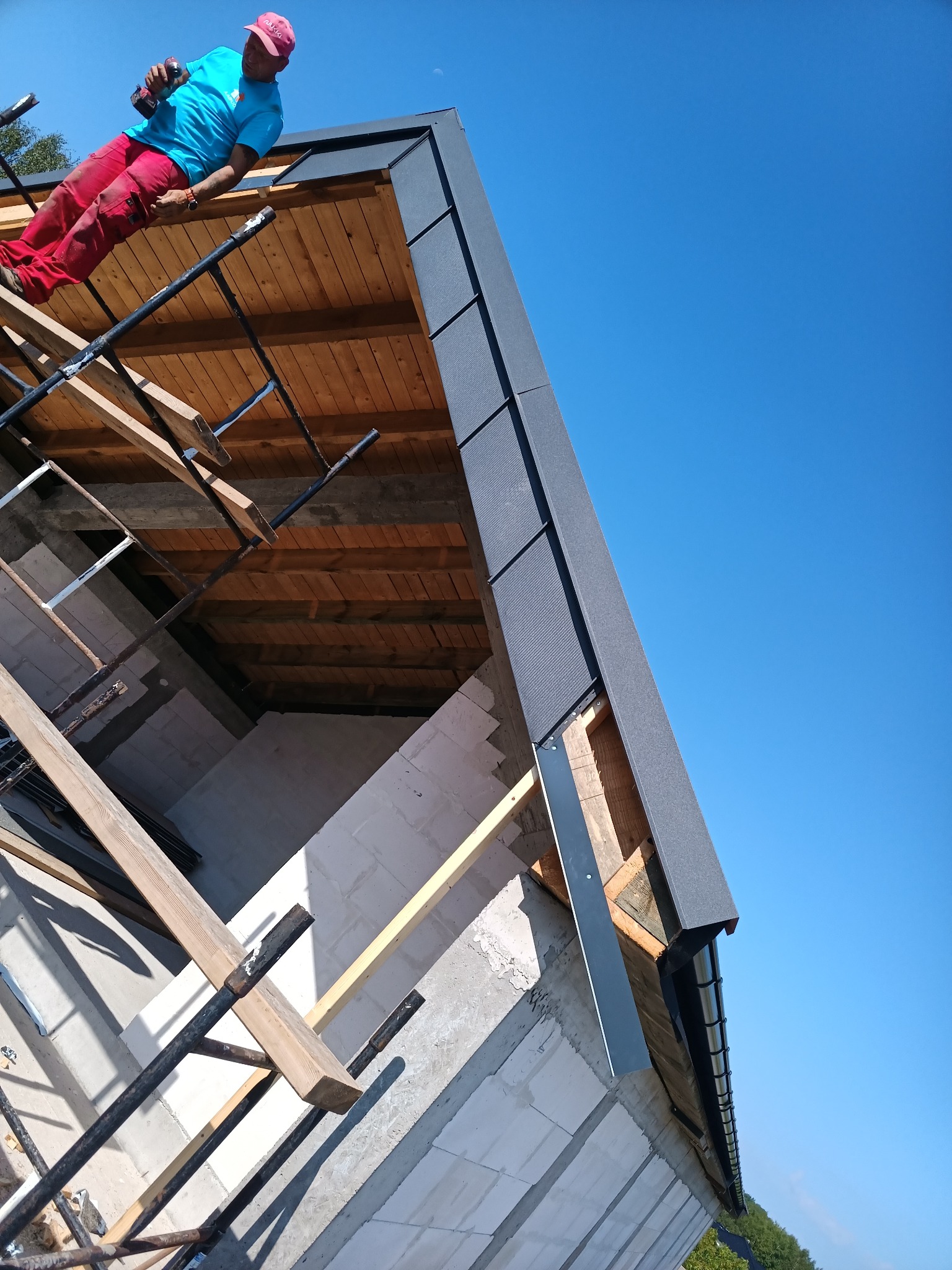 Pracownik w czerwonych spodniach montuje szarą blachodachówkę na drewnianej konstrukcji dachu domu w trakcie budowy, widoczne rusztowania i nieotynkowane ściany z białego bloczka.