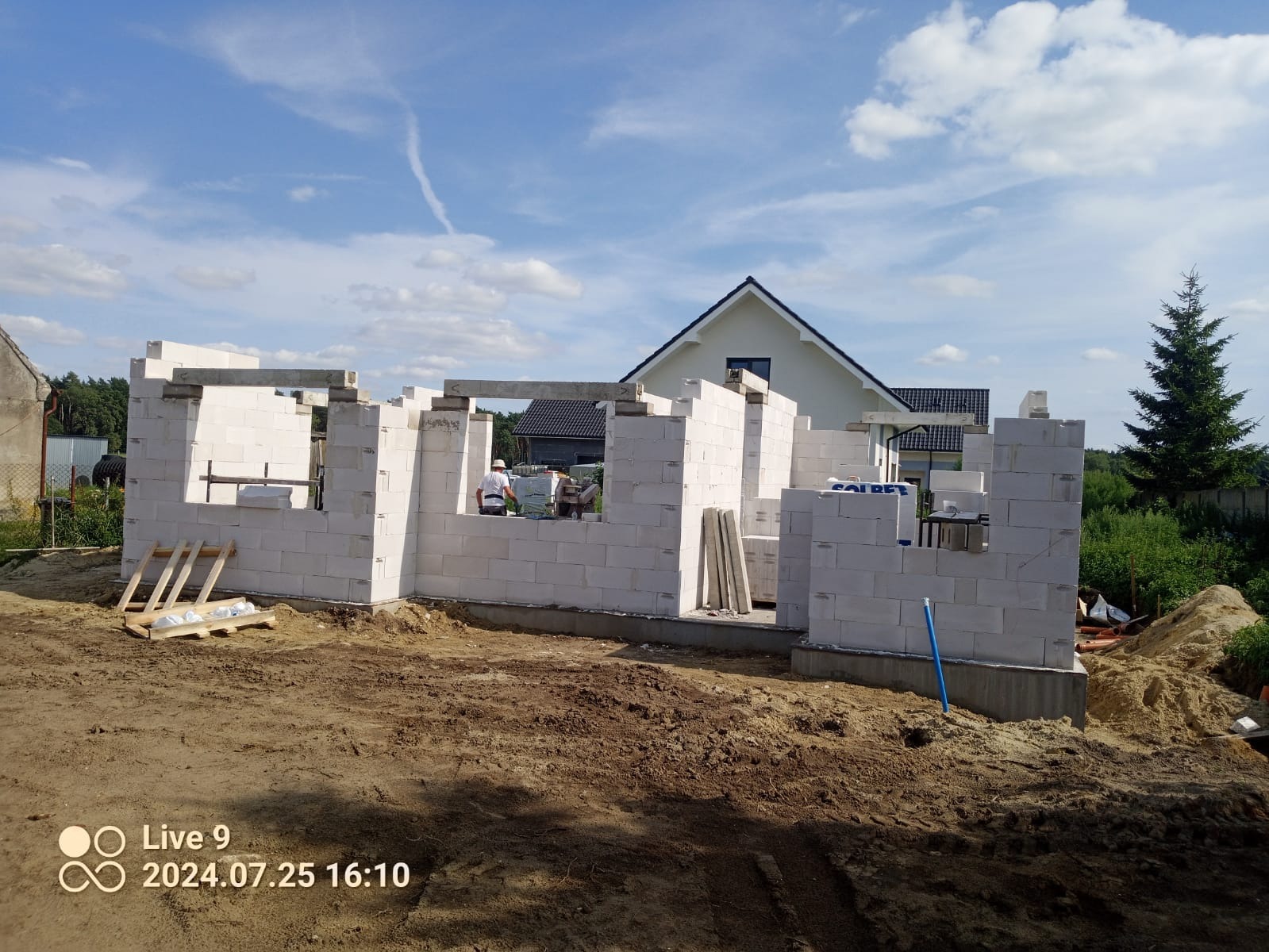 Wznoszenie ścian parteru z białych bloczków betonu komórkowego na fundamencie, widoczny pracownik w kasku, w tle dom jednorodzinny i błękitne niebo.