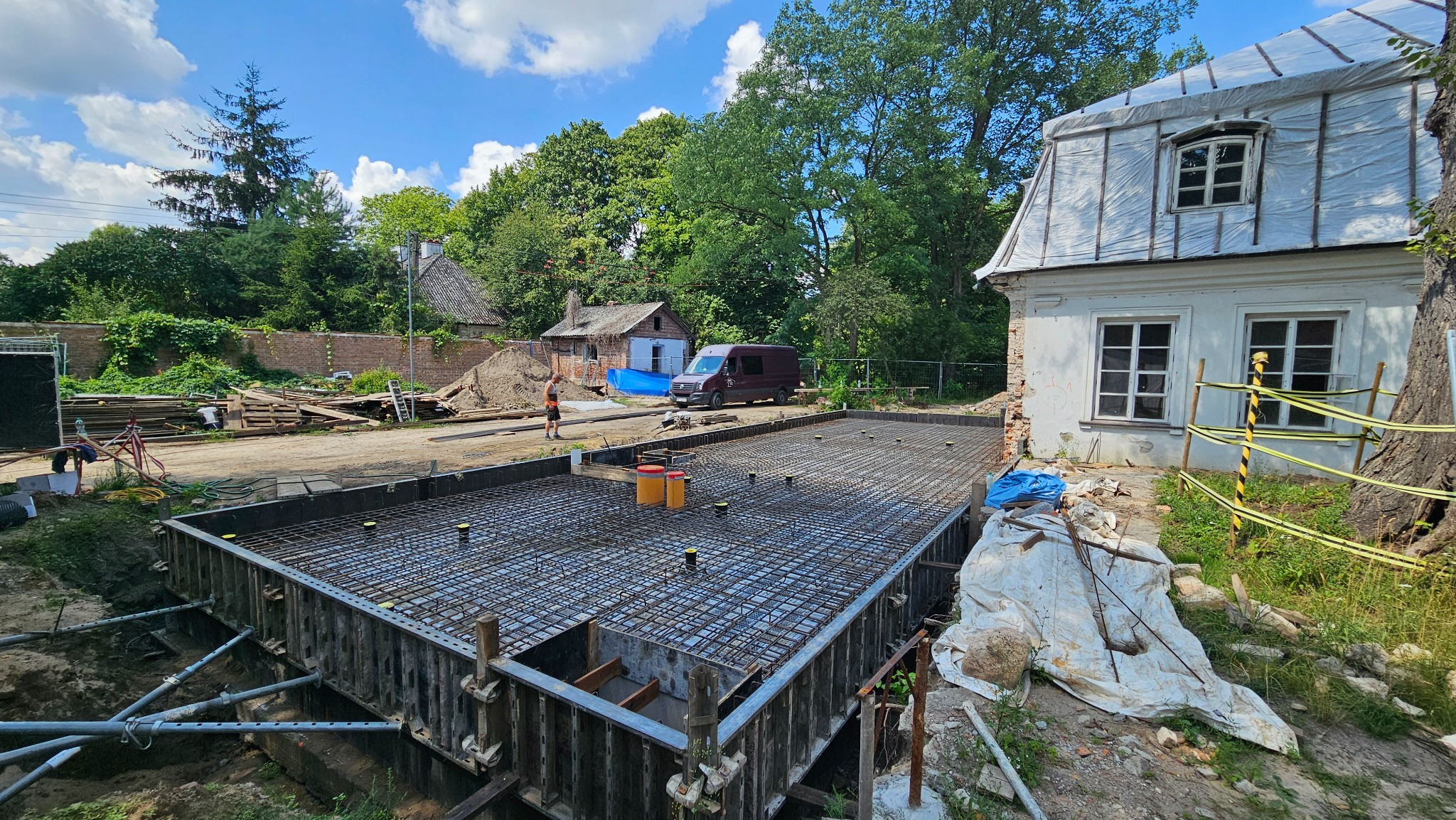 Zbrojenie fundamentu pod budynek z widocznymi żółtymi elementami dystansowymi, szalunek stalowy, budynek w tle zabezpieczony folią, pracownik w pomarańczowej koszulce.