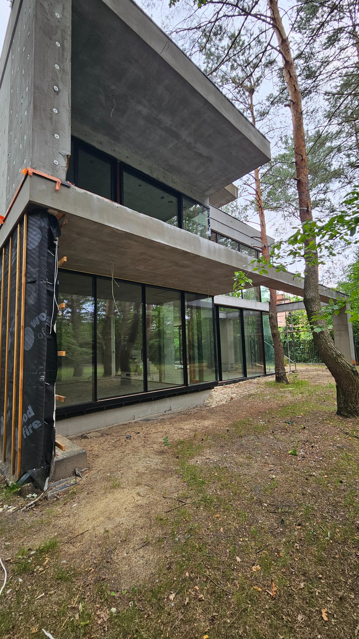 Surowy betonowy dom w stylu modernistycznym w trakcie budowy, z dużymi oknami i widoczną izolacją termiczną na elewacji, otoczony drzewami.