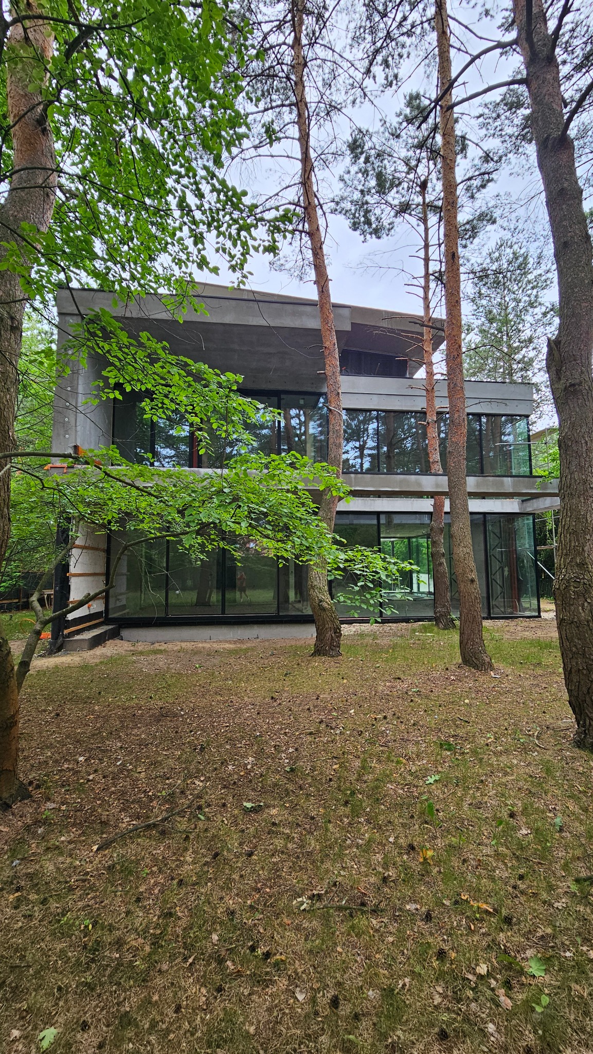 Nowoczesny dom z betonu architektonicznego z dużymi przeszkleniami, otoczony drzewami iglastymi i liściastymi, widoczny z zewnątrz na tle pochmurnego nieba.