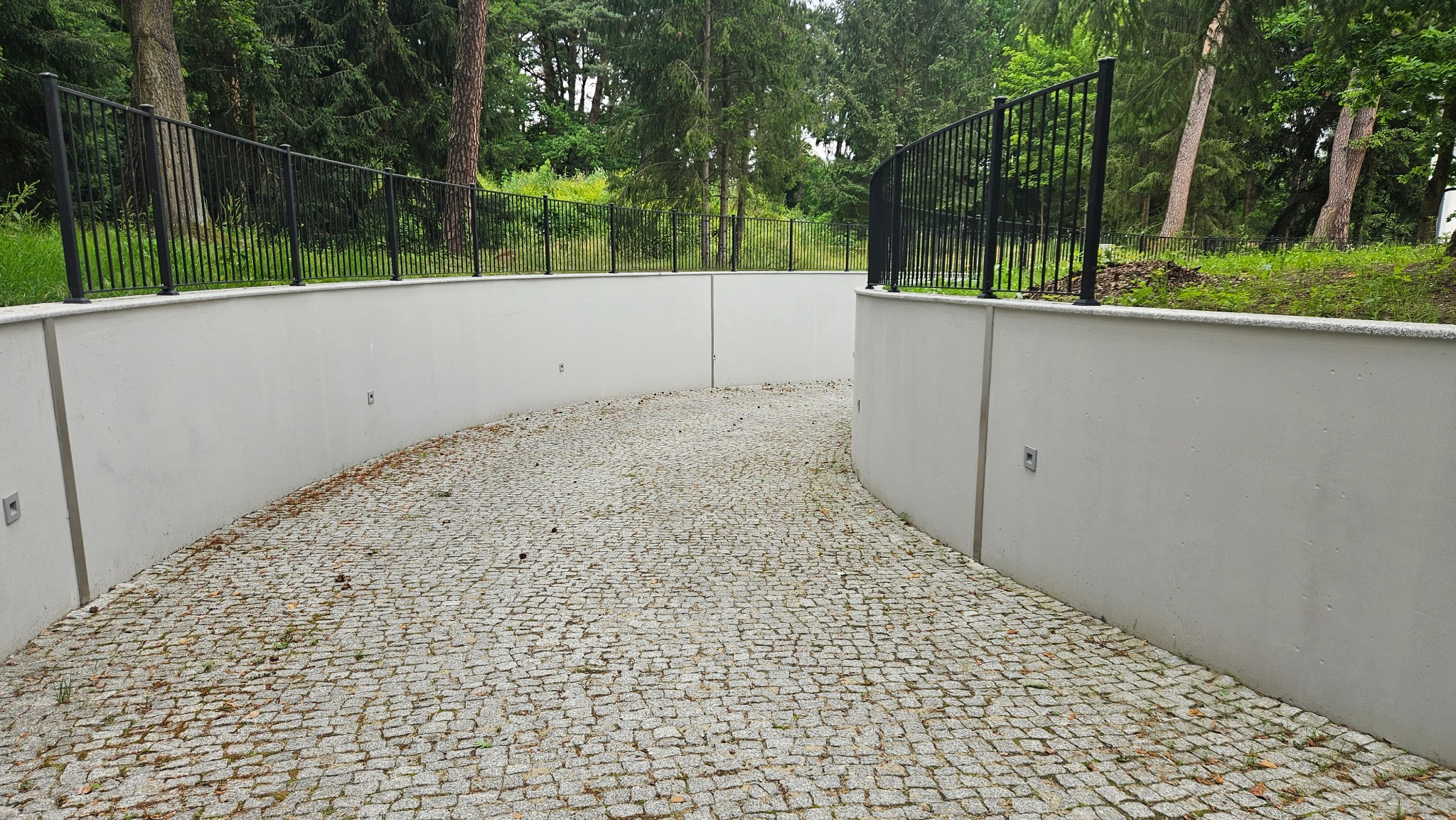 Betonowy mur oporowy z czarnym metalowym ogrodzeniem i kostką brukową w Piasecznie, otoczony zielenią.