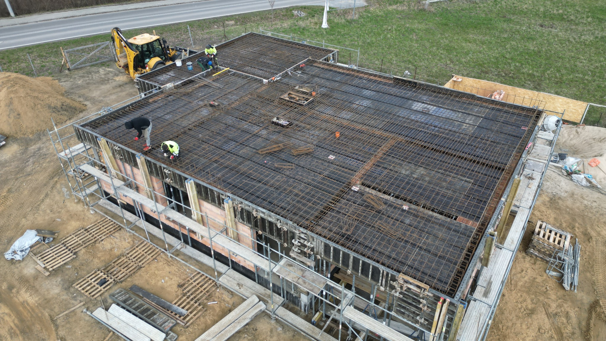 Zbrojenie stropu budynku jednorodzinnego, widok z góry na siatkę zbrojeniową, szalunki, rusztowania i pracujących robotników w kaskach, z tyłu koparka.