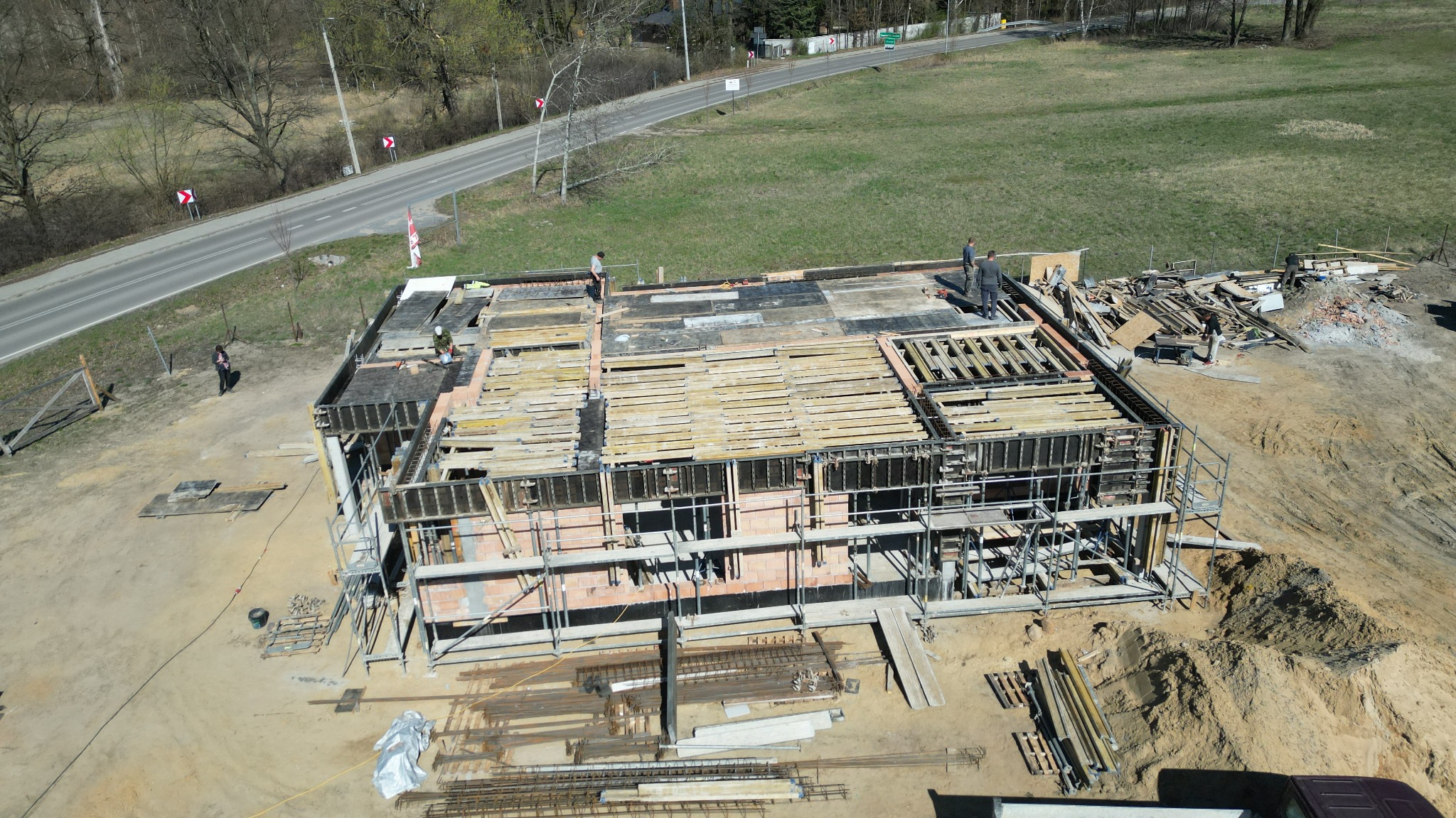 Budowa domu jednorodzinnego w stanie surowym otwartym, widok z góry na szalunki i zbrojenia, w tle droga i drzewa, kilka osób na budowie.